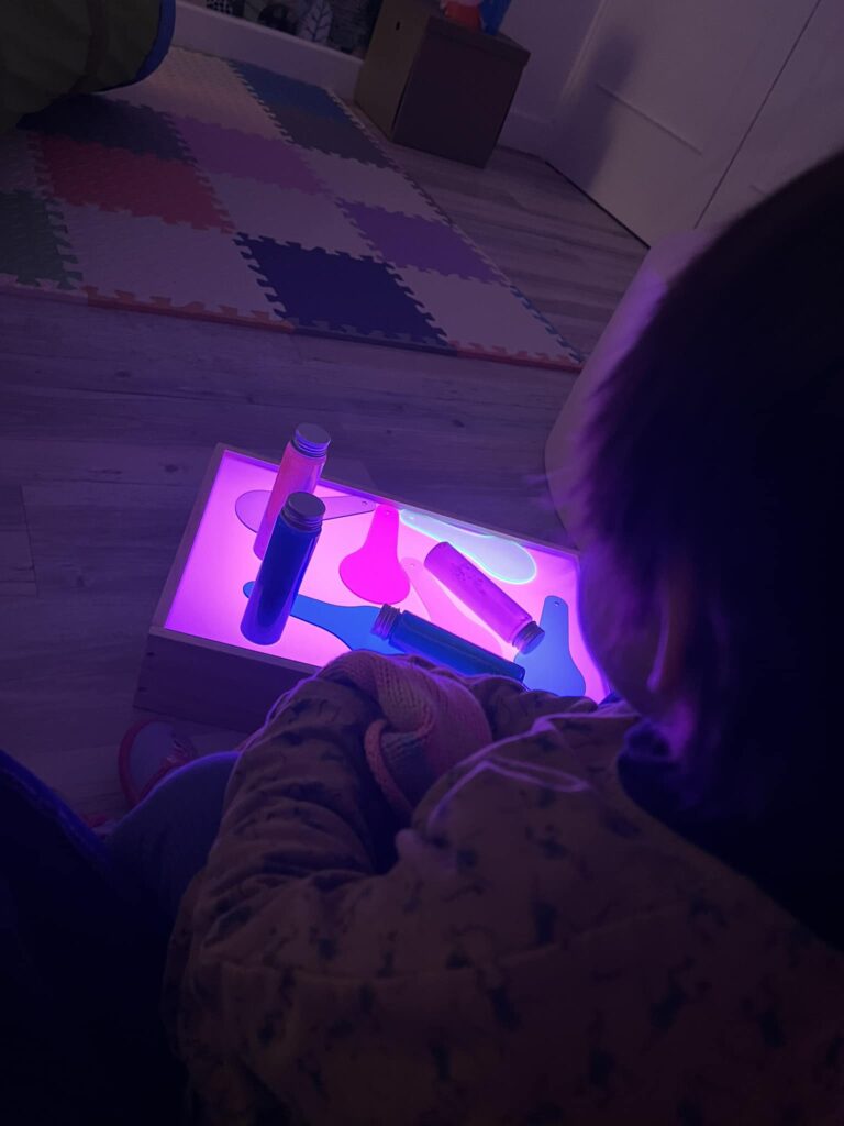 Enfant vu de dos, jouant avec une table lumineuse illuminée en violet sur laquelle sont posés des objets colorés et transparents. Illustration de l'activité d'éveil sensoriel et lumineux à la MAM Super Poppins à Blagnac