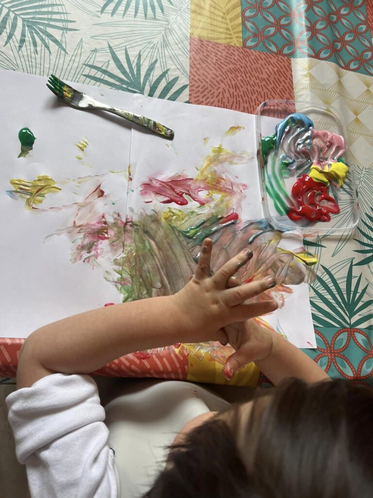  Vue de haut d'un enfant en train de peindre avec ses doigts et une fourchette sur une feuille blanche, avec des pots de peinture colorée à côté. Activité d'éveil artistique et sensoriel à la MAM Super Poppins à Blagnac